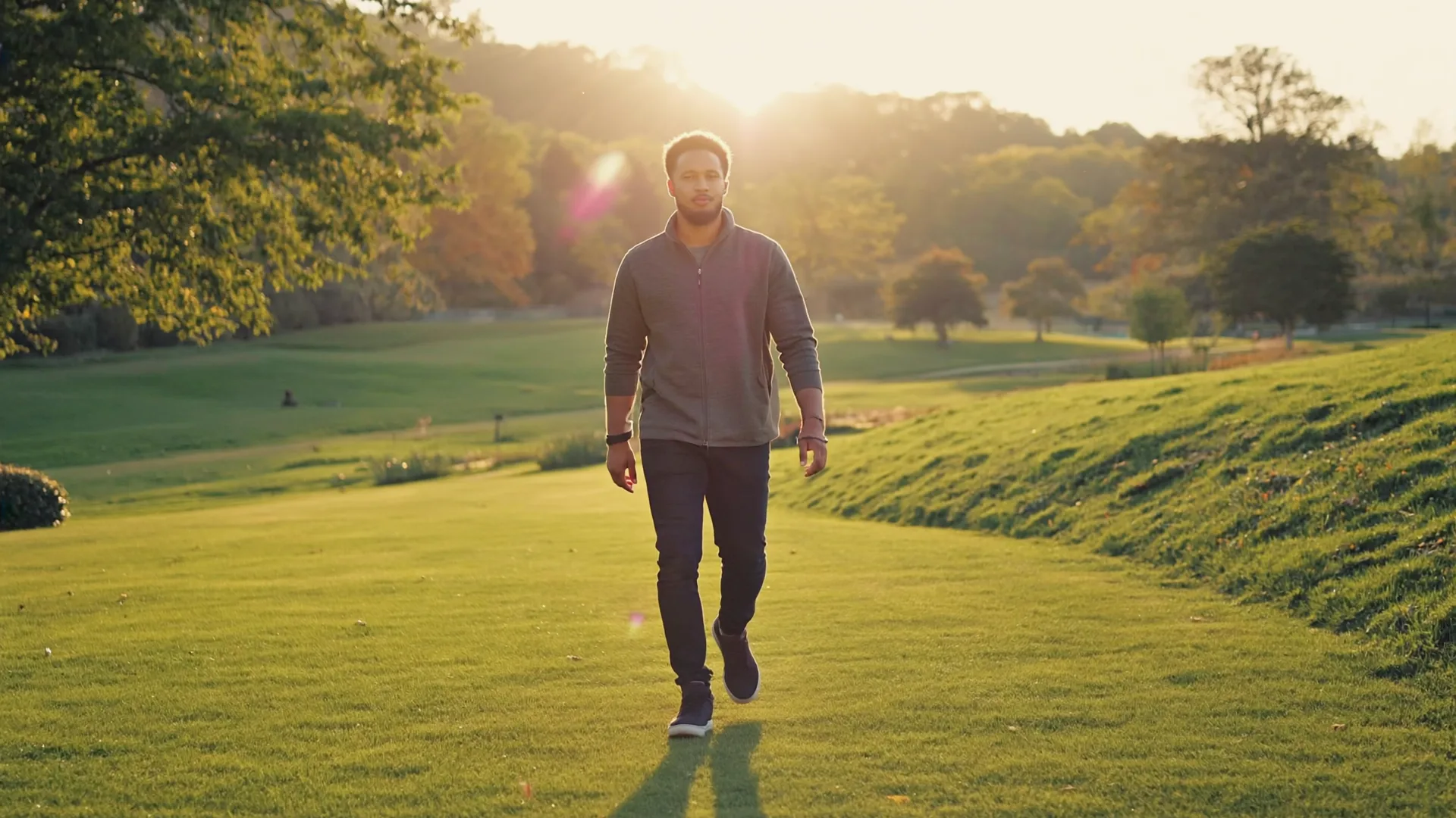 A man walking comfortably through a green park at golden hour, rolling parkland behind him
