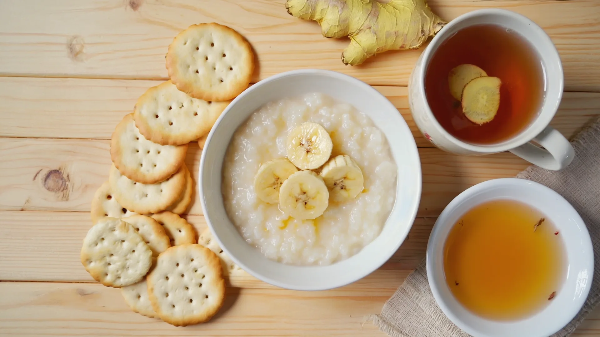 Gentle easy-to-digest foods laid out on a wooden table: porridge with banana, ginger tea, crackers and clear soup