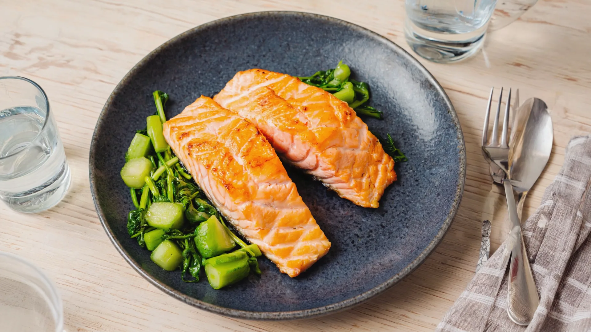 A plate of baked salmon with green vegetables and a glass of water on a wooden kitchen table