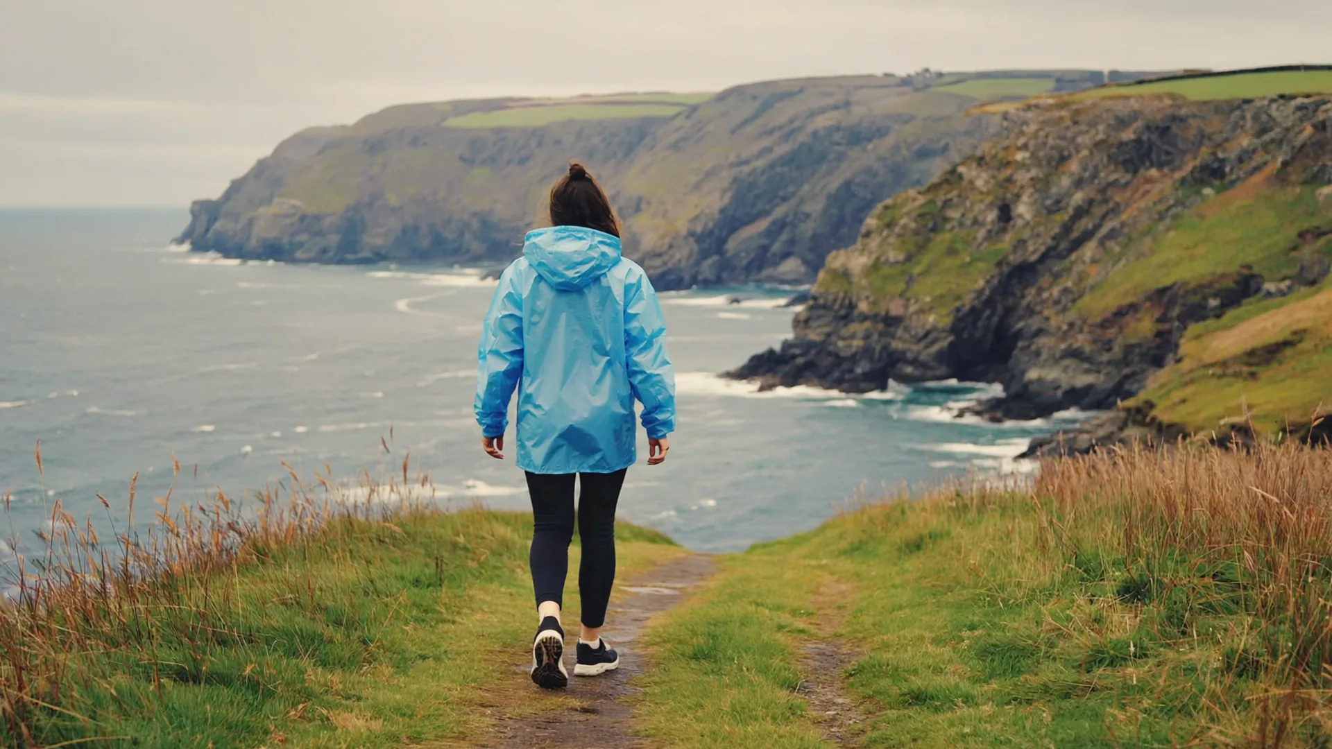 A person in a rain jacket and walking shoes walking along a green Irish coastal path beside the sea on a soft overcast day